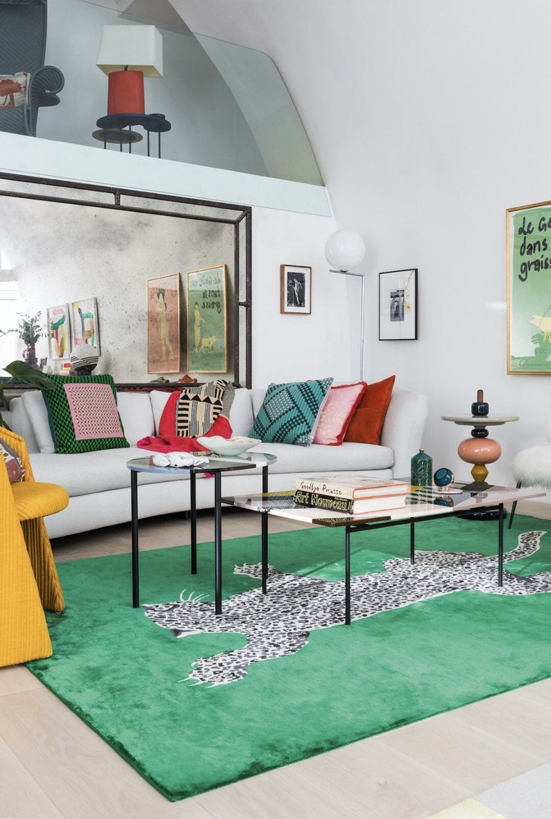 Pu&ograve; includere: Un soggiorno moderno con un divano bianco, cuscini colorati e un tappeto verde con stampa leopardata. La stanza comprende tavolini con piano in vetro, opere d'arte e una sedia gialla. Una lampada decorativa &egrave; su un tavolino.