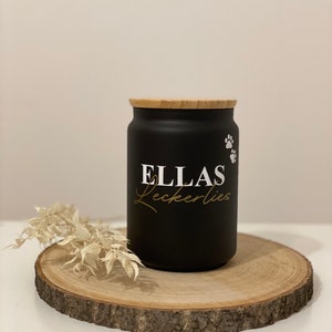 Könnte beinhalten: Schwarze Keramikdose mit Bambusdeckel, beschriftet mit "ELLAS Leckerlies" in weißer Schrift mit goldener Umrandung. Die Dose hat ein Pfotenabdruck-Design an der Seite.