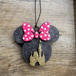 May include: A black felt Minnie Mouse head with a pink polka dot bow and a gold glitter castle. The ornament hangs from a black string. The background is a wooden surface.