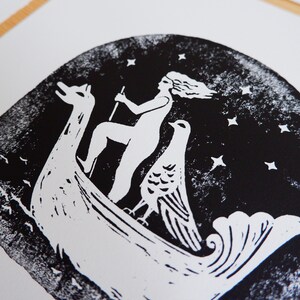 Peut inclure: Gravure sur linol&eacute;um en noir et blanc d'une femme chevauchant un bateau avec un oiseau. La femme a les cheveux longs et tient un b&acirc;ton. Le bateau a la forme d'un cygne et a un oiseau &agrave; l'arri&egrave;re. L'arri&egrave;re-plan est bleu fonc&eacute; avec des &eacute;toiles blanches.