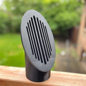 May include: A black plastic oval-shaped vent with vertical slats. The vent is mounted on a wooden surface.
