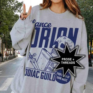 May include: A gray sweatshirt with a blue graphic print of a skull, a book, and a dagger. The text on the graphic reads "Pance Orion", "Numerology", "Zodiac Guild", "Prose + Threads", and "Professor of Dark Magic".