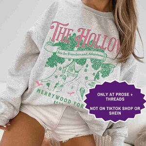 May include: A light gray sweatshirt with the text "The Hollow" in pink and green. The design includes a tree with a building and the words "Inn for Travelers and Adventurers". The sweatshirt also has the words "Merrywood For" and "Ash".