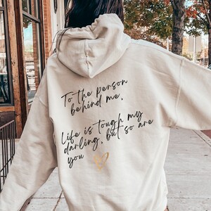 May include: Cream-colored hoodie with the text "To the person behind me. Life is tough my darling, but so are you" in black script. A small gold heart is below the text. The hoodie is worn with white high-top sneakers.