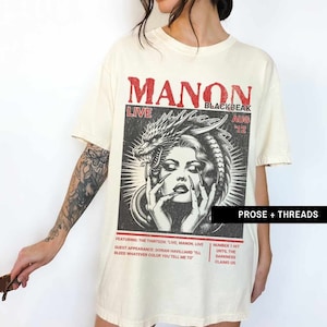 May include: An oversized white t-shirt featuring a black and white graphic print of a woman with a dragon and snake. The text on the shirt reads "MANON LIVE BLACKBEAK AUG '12" and "PROSE + THREADS". The text below the graphic reads "FEATURING: THE THIRTEEN: "LIVE, MANON. LIVE" GUEST APPEARANCE: DORIAN HAVILLIARD "I'LL BLEED WHATEVER COLOR YOU TELL ME TO" NUMBER 1 HIT UNTIL THE DARKNESS CLAIMS US"
