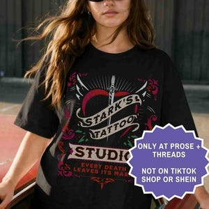 Op de afbeelding: Zwart T-shirt met de tekst "STARK'S TATTOO STUDIO" in een gestileerd ontwerp met een hart, vleugels en een dolk. Het shirt heeft ook de tekst "EVERY DEATH LEAVES ITS MARK". Het shirt wordt gedragen met een wit-blauwe pet.