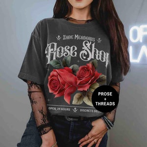 May include: Black t-shirt with a vintage-style graphic of red roses and the text "Zade Meadows Rose Shop" with skull and crossbones. The text "Open 24 Hours" and "Discrete Delivery" is also visible on the shirt.