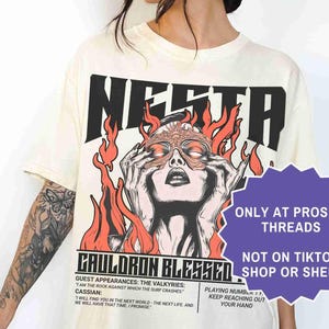 May include: Cream-colored graphic t-shirt featuring the word "NESTA" in black block letters. The shirt has a graphic of a woman with flames and the text "CAULDRON BLESSED". Additional text includes "ONLY AT PROSE + THREADS" and "NOT ON TIKTOK SHOP OR SHEIN".