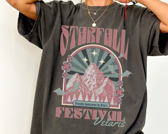 May include: A dark gray oversized t-shirt with a vintage graphic print of a mountain range with stars and bats. The text on the shirt reads "Starfall Festival Velaris" and "Proudly Sponsored by Rita's".