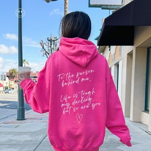 May include: A bright pink hoodie with white text on the back. The text reads, "To the person behind me, Life is tough my darling, but so are you" with a heart symbol. The hoodie is worn by a person walking on a sidewalk.