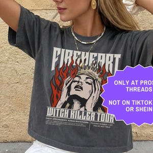 May include: A charcoal grey t-shirt featuring the text "FIREHEART" and an illustration of a woman wearing a crown. Below is "WITCH KILLER TOUR". The shirt is worn with a white skirt. The image also contains text that says "ONLY AT PROSE + THREADS NOT ON TIKTOK SHOP OR SHEIN".