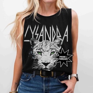 May include: Black sleeveless tank top with the word "LYSANDRA" in white block letters. The shirt features a detailed black and white leopard face with green eyes. The text "PROSE + THREADS" is also visible.