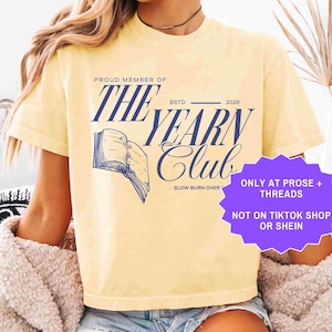 May include: A pale yellow t-shirt with navy blue text reading "PROUD MEMBER OF THE YEARN CLUB". The design includes an open book graphic and the words "SLOW BURN OVER". A purple sticker states "ONLY AT PROSE + THREADS NOT ON TIKTOK SHOP OR SHEIN".