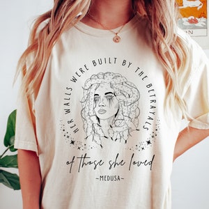 May include: Cream-colored t-shirt featuring a black and white illustration of Medusa with the text "HER WALLS WERE BUILT BY THE BETRAYALS of those she loved - MEDUSA-". The design is encircled by stars.
