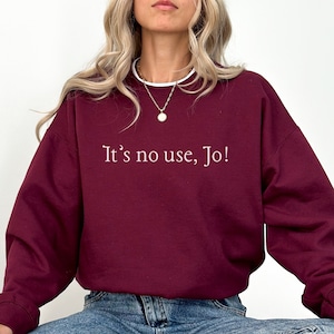 May include: A maroon sweatshirt with the text "It's no use, Jo!" printed in white on the front. The model is wearing blue jeans and black trainers.