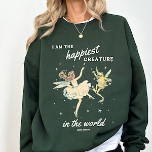 May include: Green sweatshirt with a white graphic print of a fairy and a frog dancing. The text on the sweatshirt reads "I am the happiest creature in the world. Jane Austen."