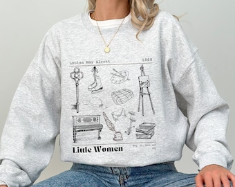 Könnte beinhalten: Ein graues Sweatshirt mit einer schwarz-weißen Illustration eines Buches, eines Schlüssels, eines Klaviers, eines Pinsels, eines Hutes, einer Feder und anderer Gegenstände. Der Text "Louisa May Alcott 1868" und "Little Women" ist auf dem Sweatshirt aufgedruckt.
