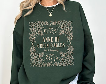 May include: A dark green sweatshirt with a white graphic print of the book cover for "Anne of Green Gables" by Lucy M. Montgomery. The print features a floral border and the title in a cream color.