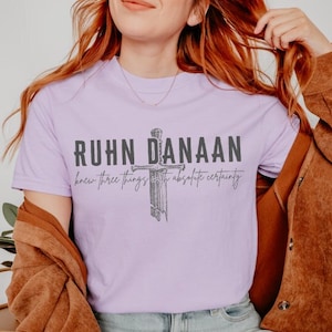 May include: A lavender t-shirt featuring a graphic of a sword and the text "Ruhn Danaan knew three things with absolute certainty".