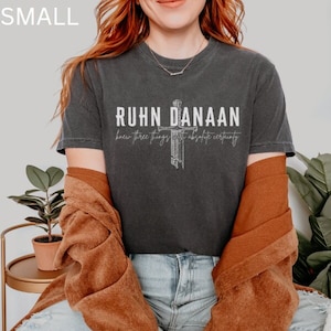 May include: A dark grey t-shirt with a white graphic design that says "RUHN DANAAN knew three things with absolute certainty". The graphic design features a stylised cross.