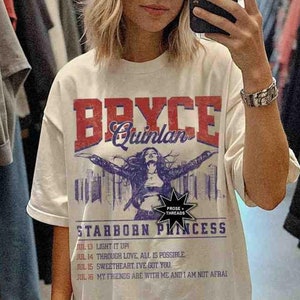 May include: A white t-shirt featuring a graphic print of a woman with the text "Bryce Quinlan" and "Starborn Princess". The shirt also includes lyrics from a song with the dates and titles of each line.