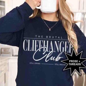 Puede incluir: Sudadera de manga larga azul marino con el texto "The Brutal Cliffhanger Club" en blanco. Los textos "Still Crying" y "Still Waiting" est&aacute;n impresos debajo del texto principal. Una estrella negra con el texto "Prose + Threads" est&aacute; impresa en el lado derecho de la sudadera.