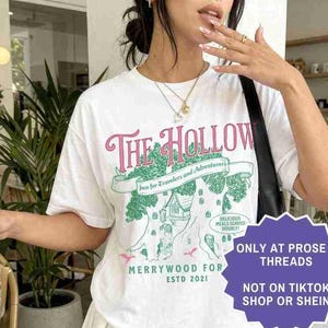 Pu&ograve; includere: T-shirt bianco con grafica rosa e verde con la scritta "The Hollow", con un'illustrazione di un albero e una casa. La maglietta ha anche il testo "Merrywood For Estd 2021". &Egrave; visibile anche una gonna a pieghe bianca.