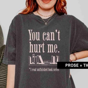 May include: A dark gray t-shirt with the text "You can't hurt me." and a graphic of a bookshelf with books. The text below the graphic says "*I read unfinished book series".