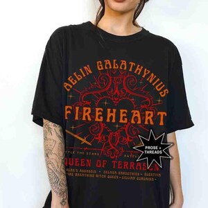 Pode incluir: T-shirt preto com o texto "AELIN GALATHYNIUS FIREHEART QUEEN OF TERRASEN" em laranja e vermelho. A camisa tem um design gr&aacute;fico com uma espada e estrelas. A camisa &eacute; usada por uma pessoa.