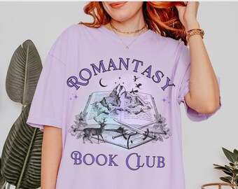 May include: A lavender t-shirt with a graphic design of a book with mountains, a deer, and stars. The text "Romantasy Book Club" is printed on the shirt.