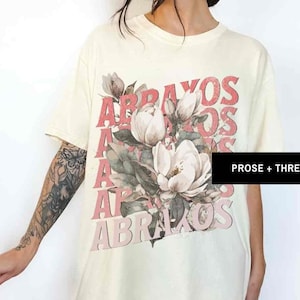Puede incluir: Una camiseta de color crema con un gráfico floral y el texto "ABRAZO" repetido en rosa. El texto está superpuesto y ligeramente desplazado. El gráfico floral presenta una gran flor de magnolia blanca con hojas verdes.