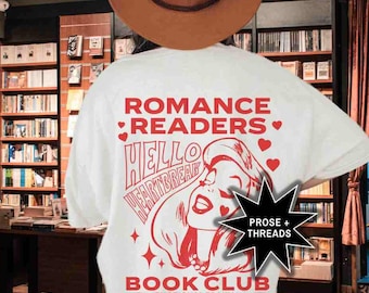 Op de afbeelding: Een off-white t-shirt met rode grafische tekst die ROMANCE READERS BOOK CLUB, HELLO HEARTBREAK en EMOTIONAL TRAUMA GUARANTEED leest. Het ontwerp bevat een gestileerde illustratie van een persoon en kleine hartpictogrammen.