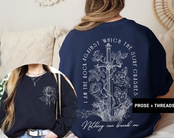 May include: Navy blue sweatshirt with a white graphic of a sword, roses, and text that reads "I AM THE ROCK AGAINST WHICH THE SURF CRASHES. Nothing can break me." A smaller image shows a black sweatshirt with a rose.