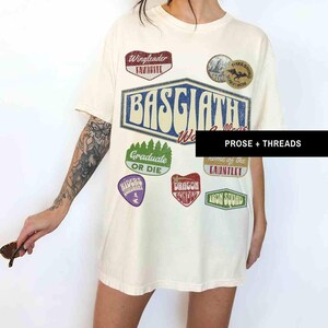 May include: A white t-shirt with a vintage-style graphic design featuring the words "Basgiath World Collective" and various patches, including "Wingleader Favorite", "Graduate or Die", "Riders Republic", "Dragon Bonder", "Home of the Gauntlet", and "Iron Squad".