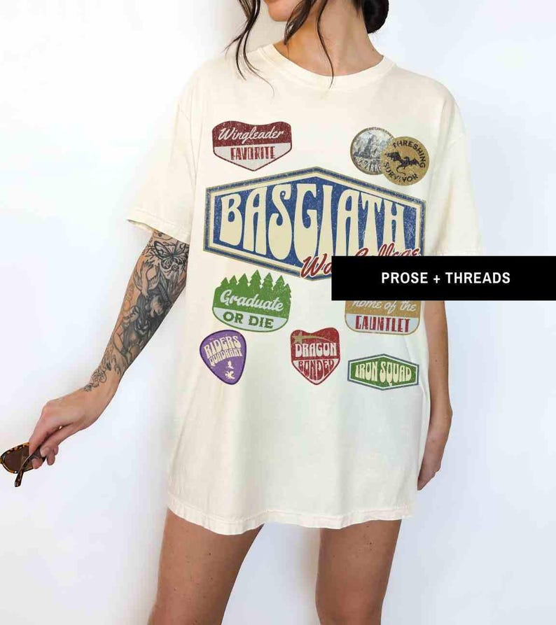 May include: A white t-shirt with a vintage-style graphic design featuring the words "Basgiath World Collective" and various patches, including "Wingleader Favorite", "Graduate or Die", "Riders Republic", "Dragon Bonder", "Home of the Gauntlet", and "Iron Squad".