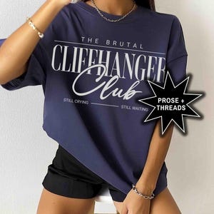 May include: A navy blue oversized t-shirt with the text "The Brutal Cliffhanger Club" in white. The text "Still Crying" and "Still Waiting" are printed below the main text. A white star with the text "Prose + Threads" is in the bottom right corner.