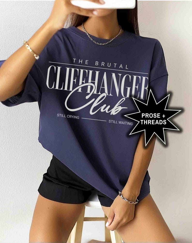 May include: A navy blue oversized t-shirt with the text "The Brutal Cliffhanger Club" in white. The text "Still Crying" and "Still Waiting" are printed below the main text. A white star with the text "Prose + Threads" is in the bottom right corner.