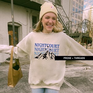 May include: A beige sweatshirt with a blue and black mountain graphic and the text "NIGHTCOURT" repeated three times. The text "PROSE + THREADS" is also visible in the bottom right corner.