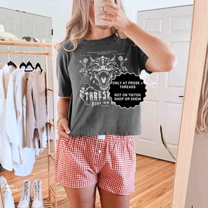 May include: A dark gray t-shirt with a dragon graphic and the text "THRESH RIDE OR". The model is wearing red and white checkered shorts. The image also includes text that says "ONLY AT PROSE + THREADS" and "NOT ON TIKTOK SHOP OR SHEIN".