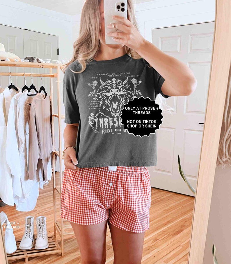 May include: A dark gray t-shirt with a dragon graphic and the text "THRESH RIDE OR". The model is wearing red and white checkered shorts. The image also includes text that says "ONLY AT PROSE + THREADS" and "NOT ON TIKTOK SHOP OR SHEIN".