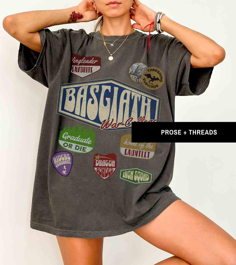 May include: A gray t-shirt with a vintage-style graphic design featuring the text "Basgaath War College" and various patches with text like "Thresing Survivor", "Graduate or Die", "Dragon Bonded", and "Iron Squad".