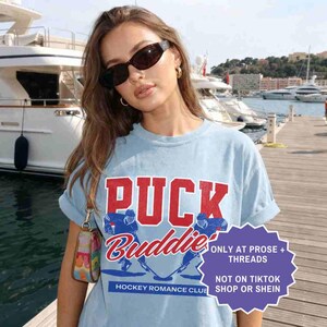 May include: Light blue t-shirt with the text "PUCK Buddies" in red and blue, featuring hockey player graphics. The shirt also has the text "HOCKEY ROMANCE CLUB" and a purple graphic with text.