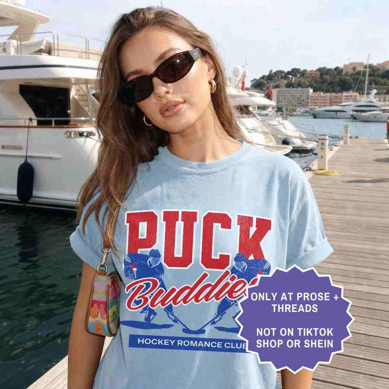 May include: Light blue t-shirt with the text "PUCK Buddies" in red and blue, featuring hockey player graphics. The shirt also has the text "HOCKEY ROMANCE CLUB" and a purple graphic with text.