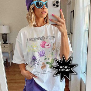 May include: A white t-shirt with a botanical illustration and the text "A Botanical Guide to Cleocal". The model is wearing a purple beanie and blue sunglasses.