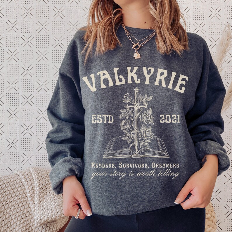 Może przedstawiać: Szara bluza z białym nadrukiem graficznym. Grafika przedstawia miecz przechodzący przez książkę z r&oacute;żami i tekst "Valkyrie EstD 2021 Readers. Survivors. Dreamers your story is worth telling".