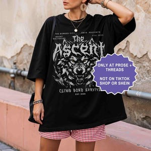 May include: Black graphic t-shirt featuring a wolf design with the text "The Ascent" and "Climb Bond Survive". Additional text includes "The Bonded City Mount Wolfsbane" and "Est. 2025". A purple sticker reads "Only at Prose + Threads" and "Not on TikTok Shop or Shein".