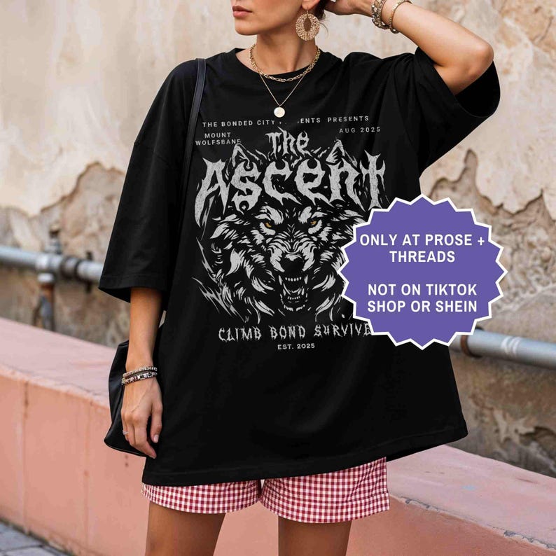 May include: Black graphic t-shirt featuring a wolf design with the text "The Ascent" and "Climb Bond Survive". Additional text includes "The Bonded City Mount Wolfsbane" and "Est. 2025". A purple sticker reads "Only at Prose + Threads" and "Not on TikTok Shop or Shein".