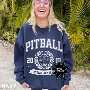 May include: Navy blue sweatshirt with white text that reads "Pitball" and "Zodiac Academy". The sweatshirt also features a white wreath with the text "Solaria Champs 2019" and a black star with the text "Prose + Threads".