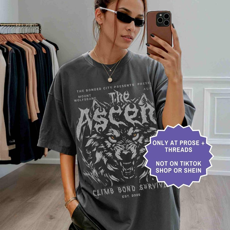 May include: Dark gray t-shirt featuring a detailed wolf graphic with the text "The Ascen". The shirt also includes the text "CLIMB BOND SURVIVE" and "EST. 2025". The shirt is available only at Prose + Threads.