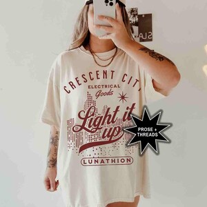May include: A white t-shirt with a red graphic design that says "Crescent City Electrical Goods Light it Up" with a star and a city skyline. The graphic also includes the text "Lunathion" and "Prose + Threads".